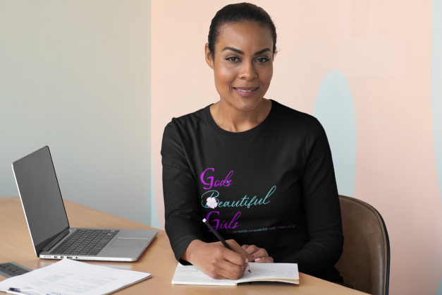 woman wearing black long sleeve shirt with the words "God's Beautiful Girls" on the front in purple and turquoise letters.  She has a laptop on the desk and she is writing in a book.