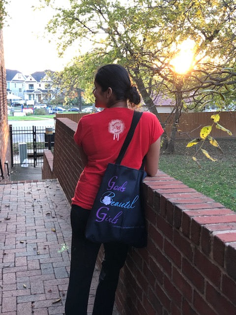 woman in red shirt with black "God's Beautiful Girls" tote bag over her shoulder. She is facing the sun and a row of houses. 