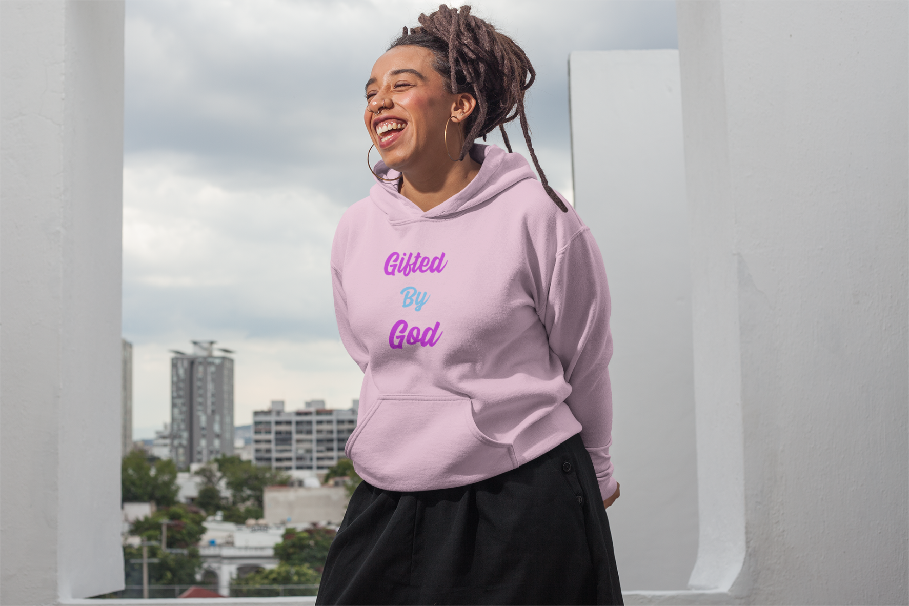 An African American Woman with locs and laughing wearing a pink pullover hoodie with the words "Gifted By God" in purple and turquoise letters.