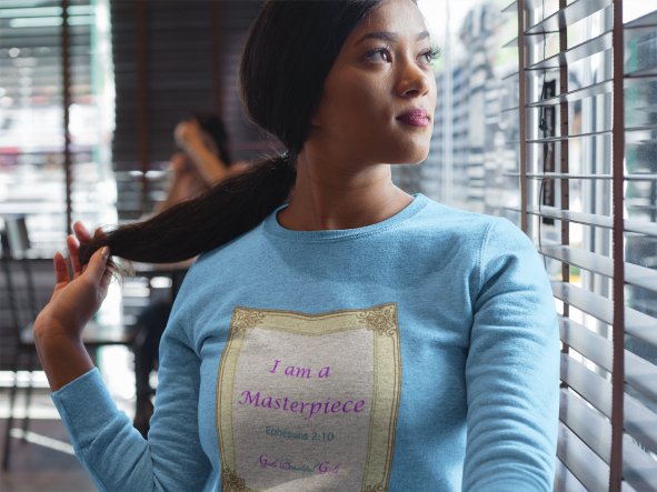 Woman wearing a light blue crewneck sweatshirt with the words "I am a Masterpiece"  on the front in a gold frame, while staring out a window.